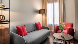 A modern living room with a grey sofa, red cushions, a red armchair, a small round table with a vase of flowers, and a floor lamp. - NH Collection León Plaza Mayor (León)