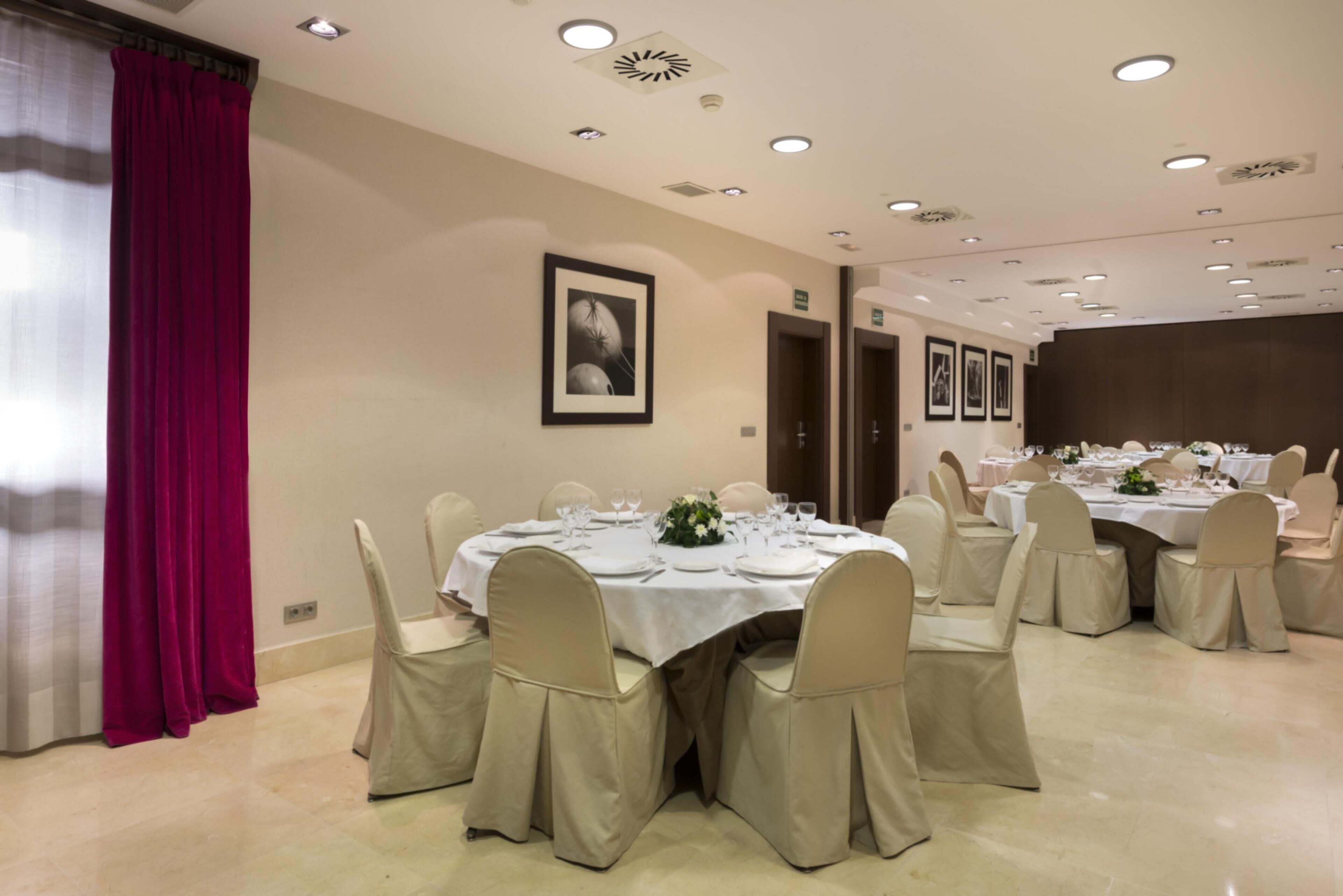 A dining area with round tables set for a meal, chairs with beige covers, and a red curtain on the left.