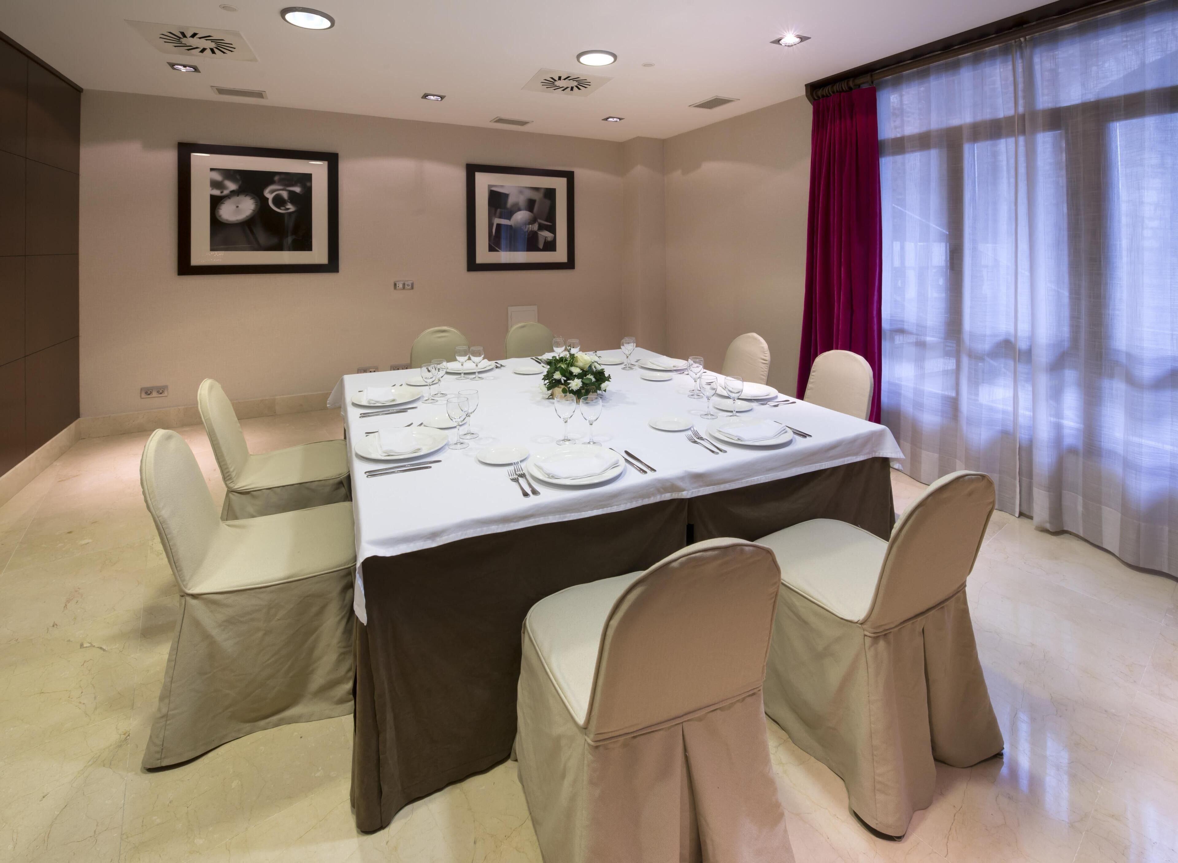 A conference room with a rectangular table set for a meeting, surrounded by chairs with white and brown covers, and two framed pictures on the wall.