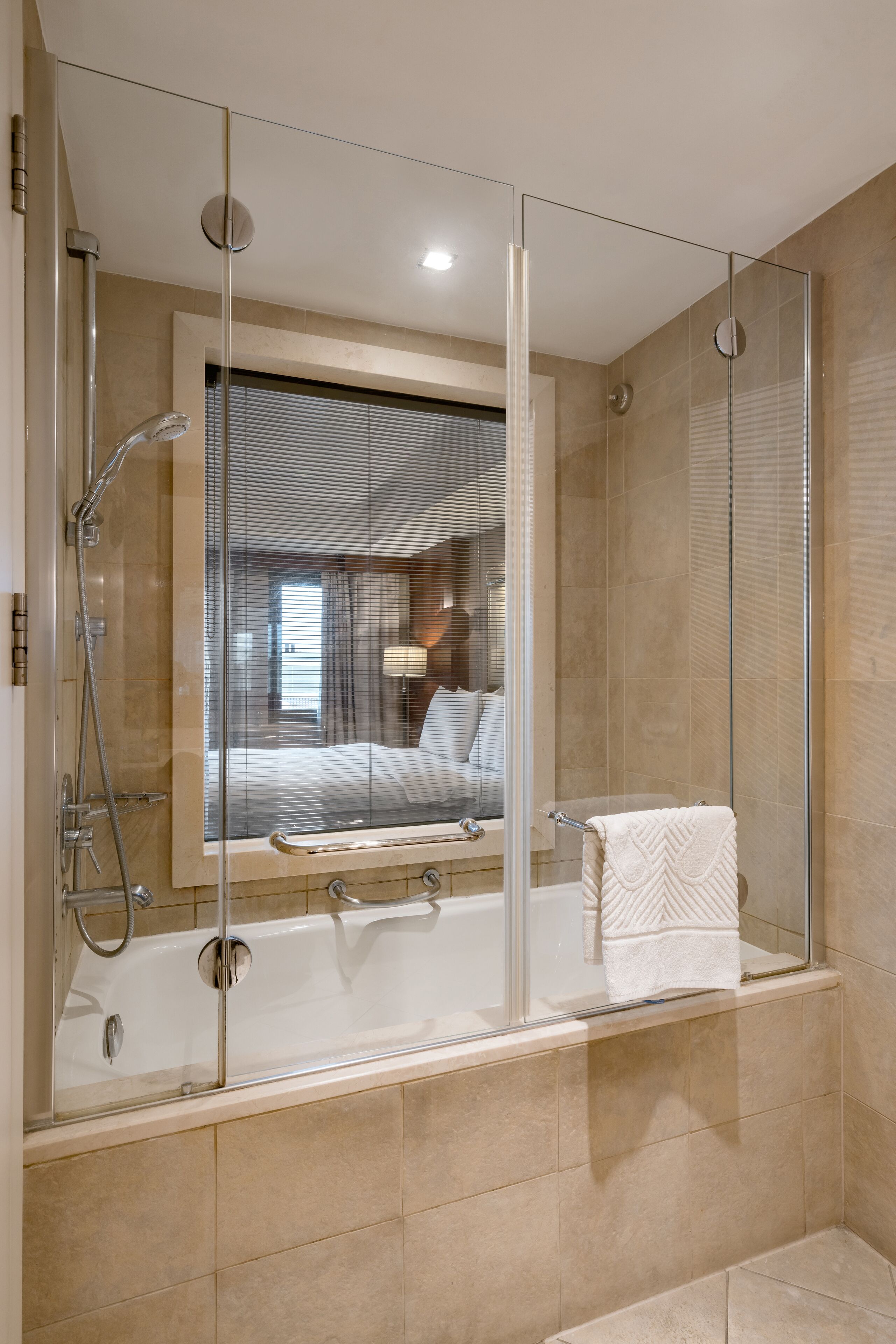 A modern bathroom with a glass-enclosed shower, a bathtub, and a visible bedroom with a bed and window.