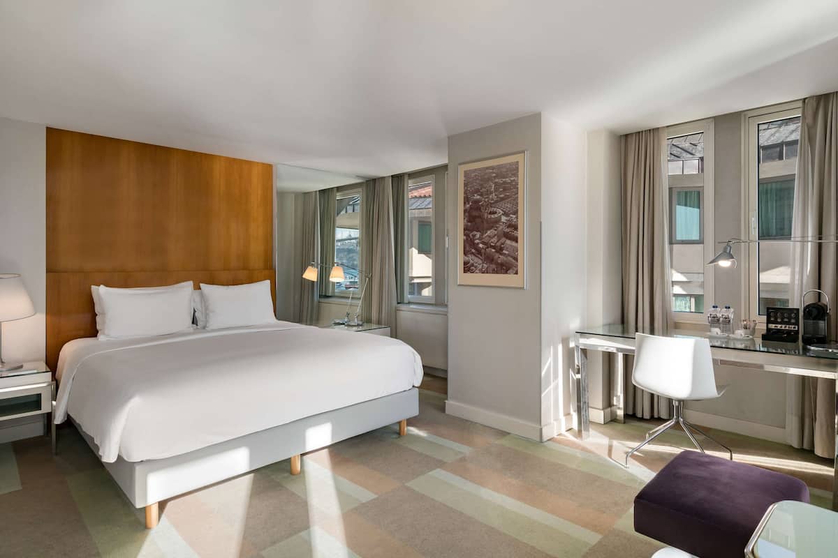 A modern hotel room with a large bed, a desk with a chair, a small table, and a window with curtains.