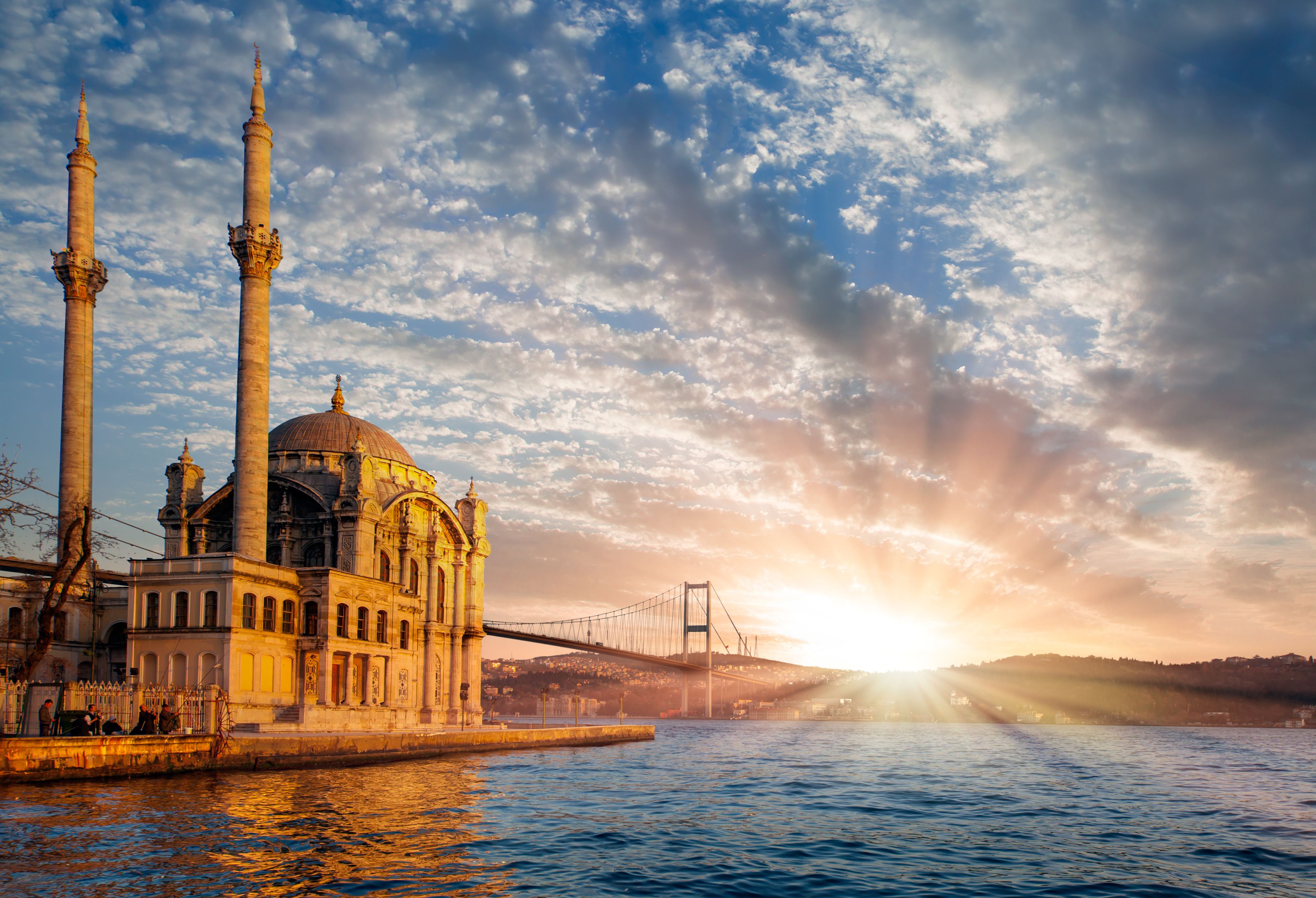 A historic building with two tall minarets and a dome, located near a body of water with a bridge in the background.