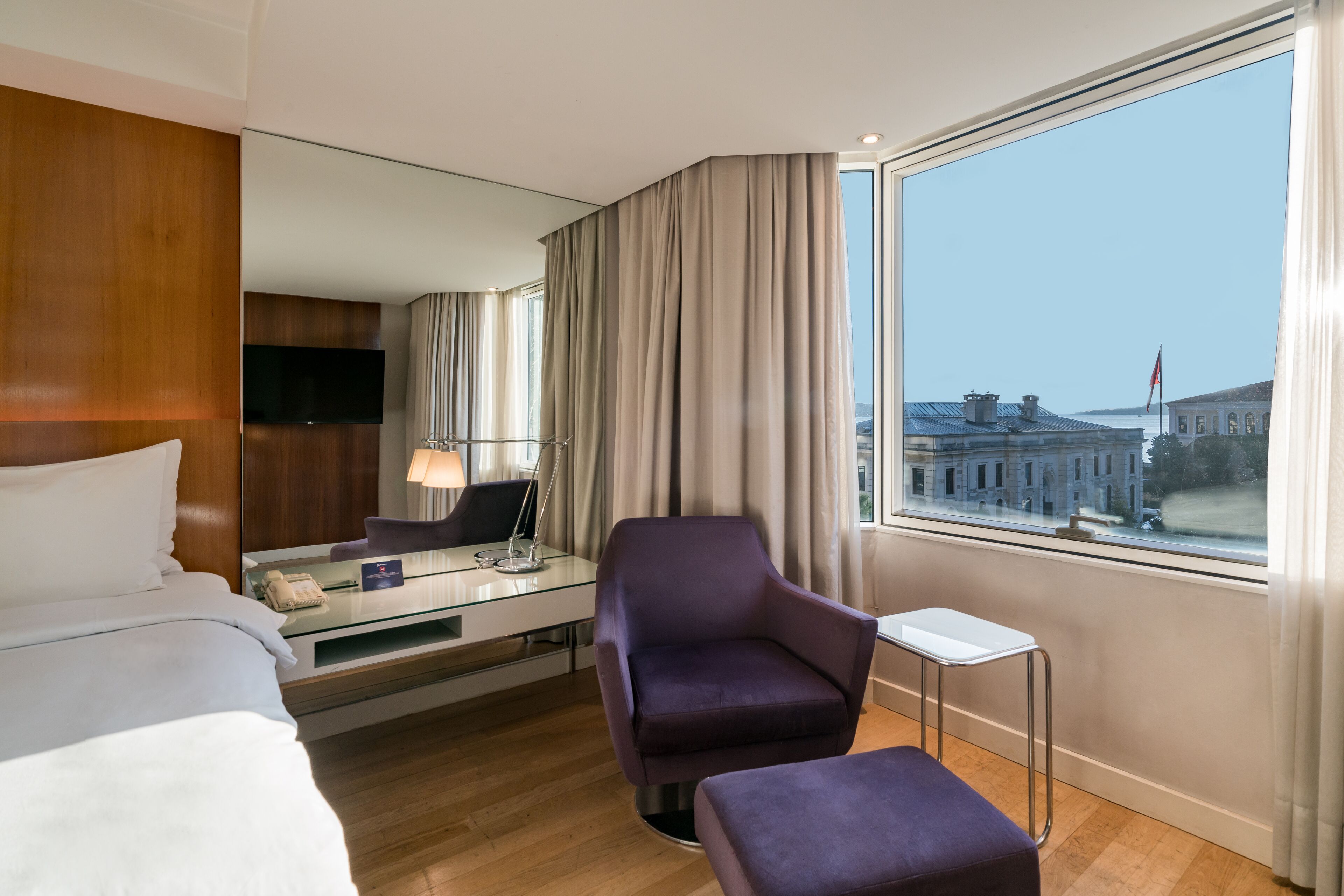 A hotel room with a bed, a desk with a lamp, a purple armchair, a small table, and a large window with a view of a building.