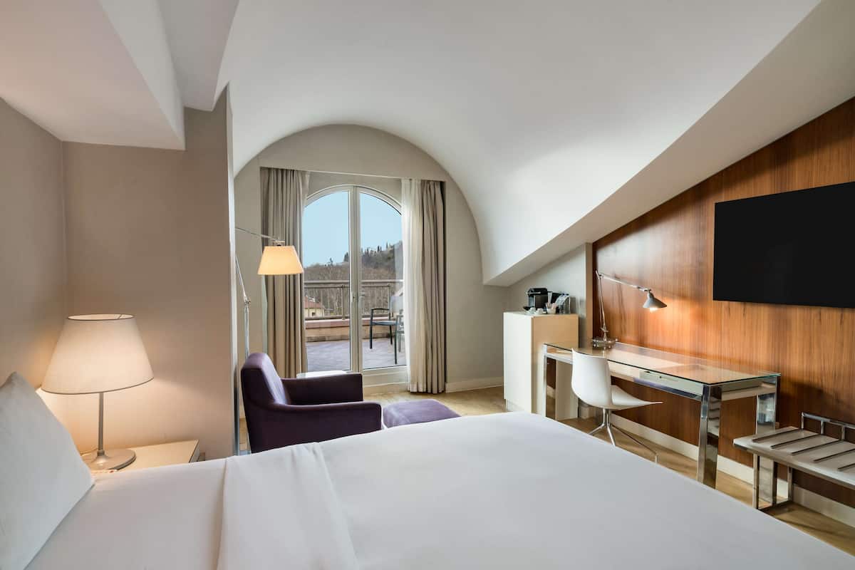 A hotel room with a bed, a desk, a chair, a TV, and a view of the outdoors.