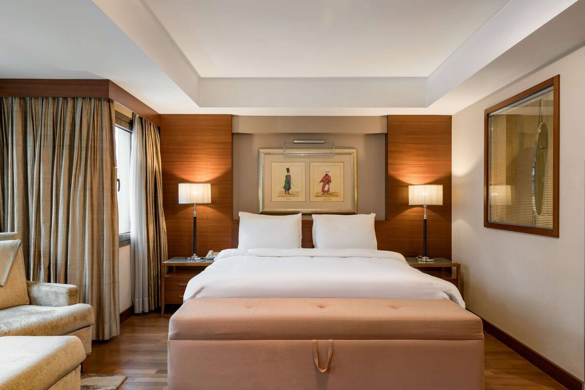 A hotel room with a large bed, two bedside tables, a desk, a chair, and a large window with curtains.