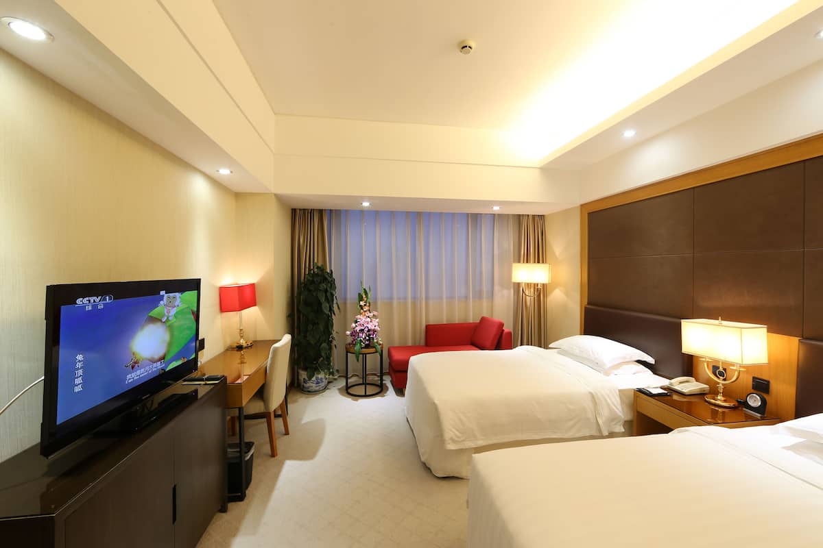 A hotel room with a television, two beds, a desk, a chair, a red sofa, a lamp, and a potted plant.