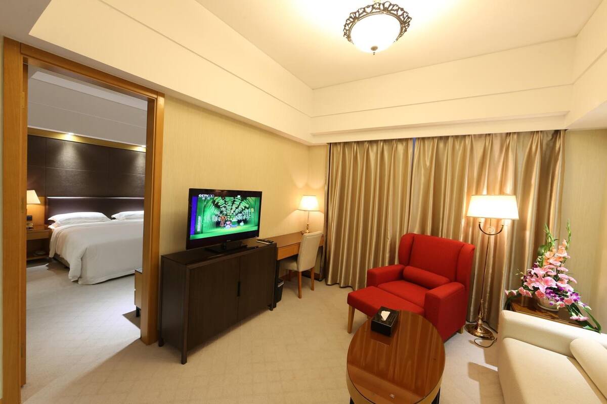 A hotel room with a bed, a TV, a red armchair, a wooden coffee table, a desk with a lamp, and a curtain.