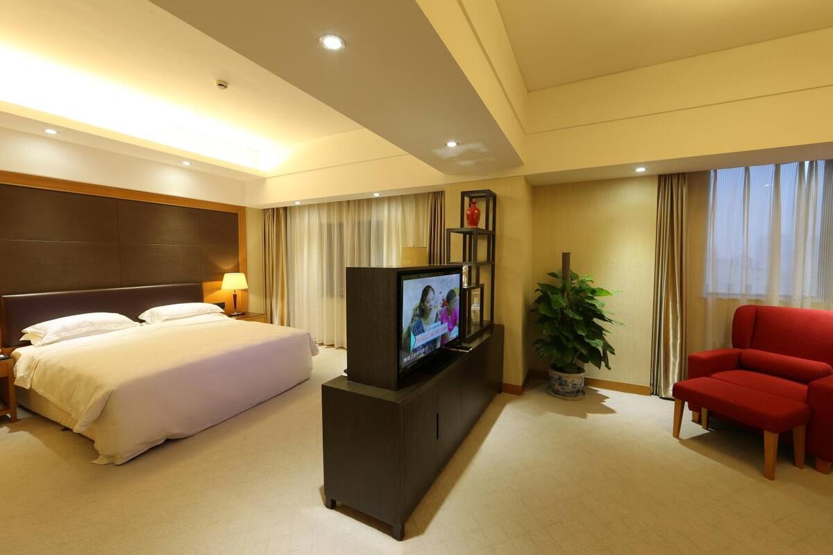 A hotel room with a bed, a TV on a stand, a red chair, and a potted plant.
