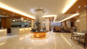 A modern hotel lobby with a central fountain, a reception desk, and a dining area with tables and chairs.