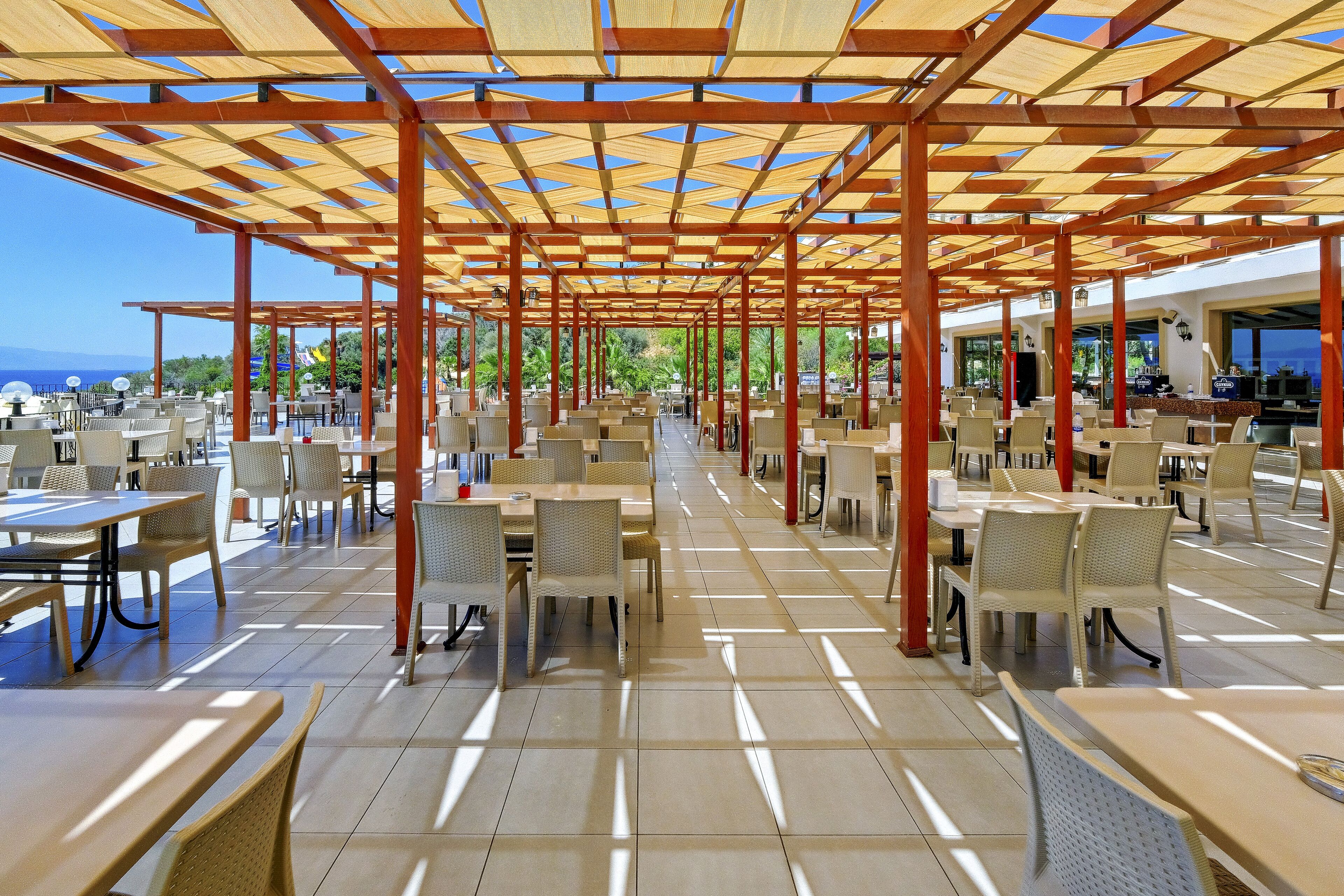 Un área de comedor al aire libre con varias mesas y sillas, piso de baldosas y una estructura de pérgola de madera.