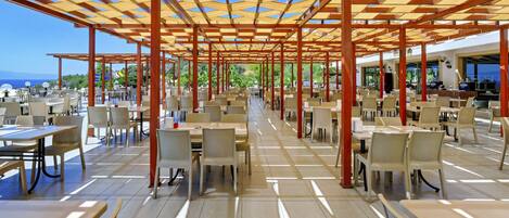 Un área de comedor al aire libre con varias mesas y sillas, piso de baldosas y una estructura de pérgola de madera.