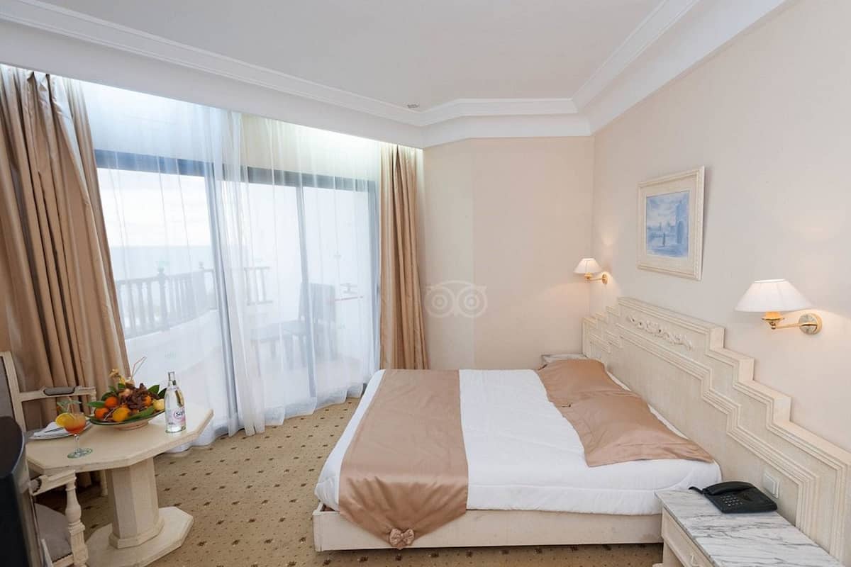 A hotel room with a bed, a desk with a fruit bowl and a drink, and a painting on the wall.