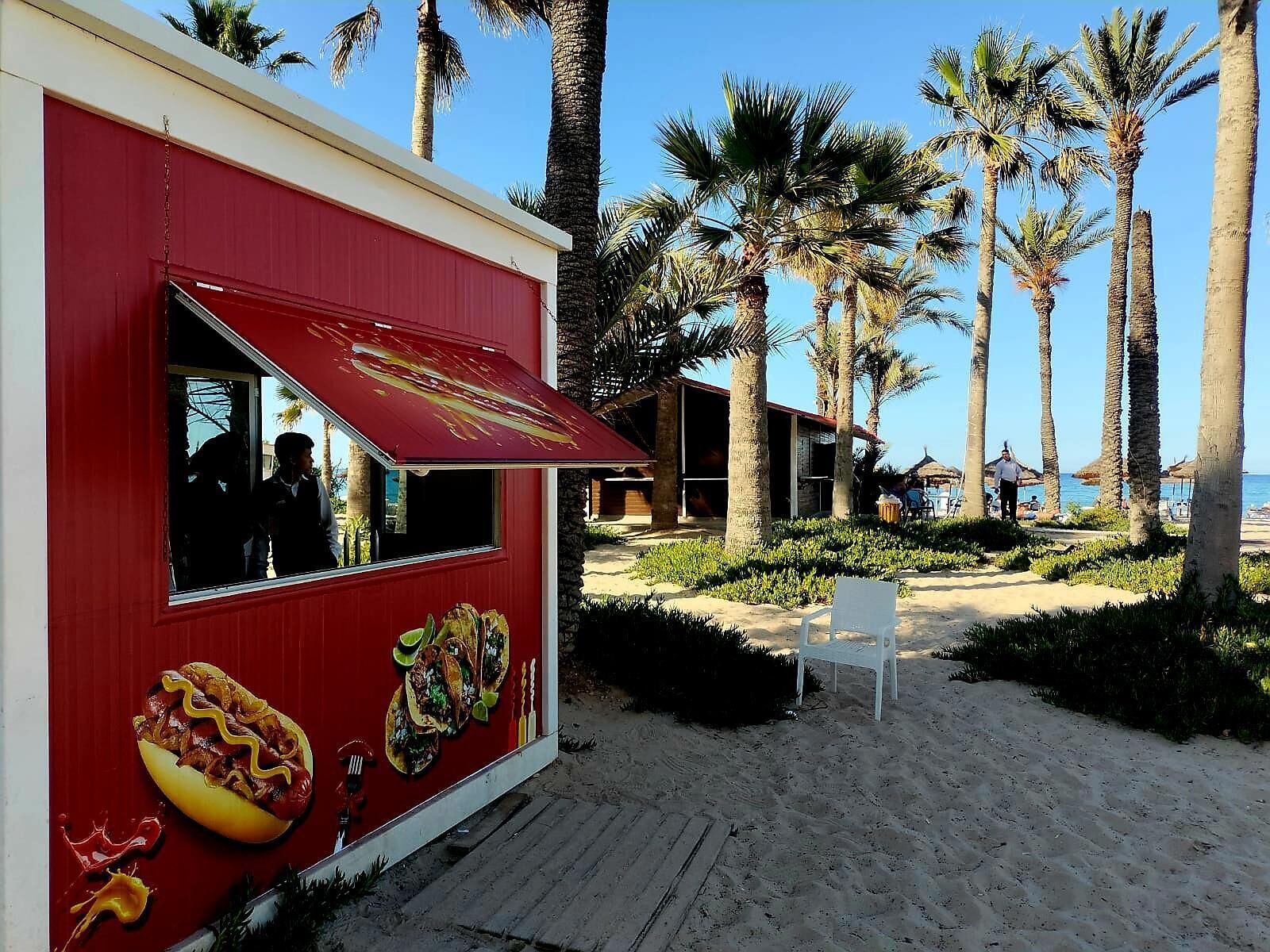 Un camión de comida rojo con un toldo grande, situado en una playa arenosa con palmeras y cielo despejado.