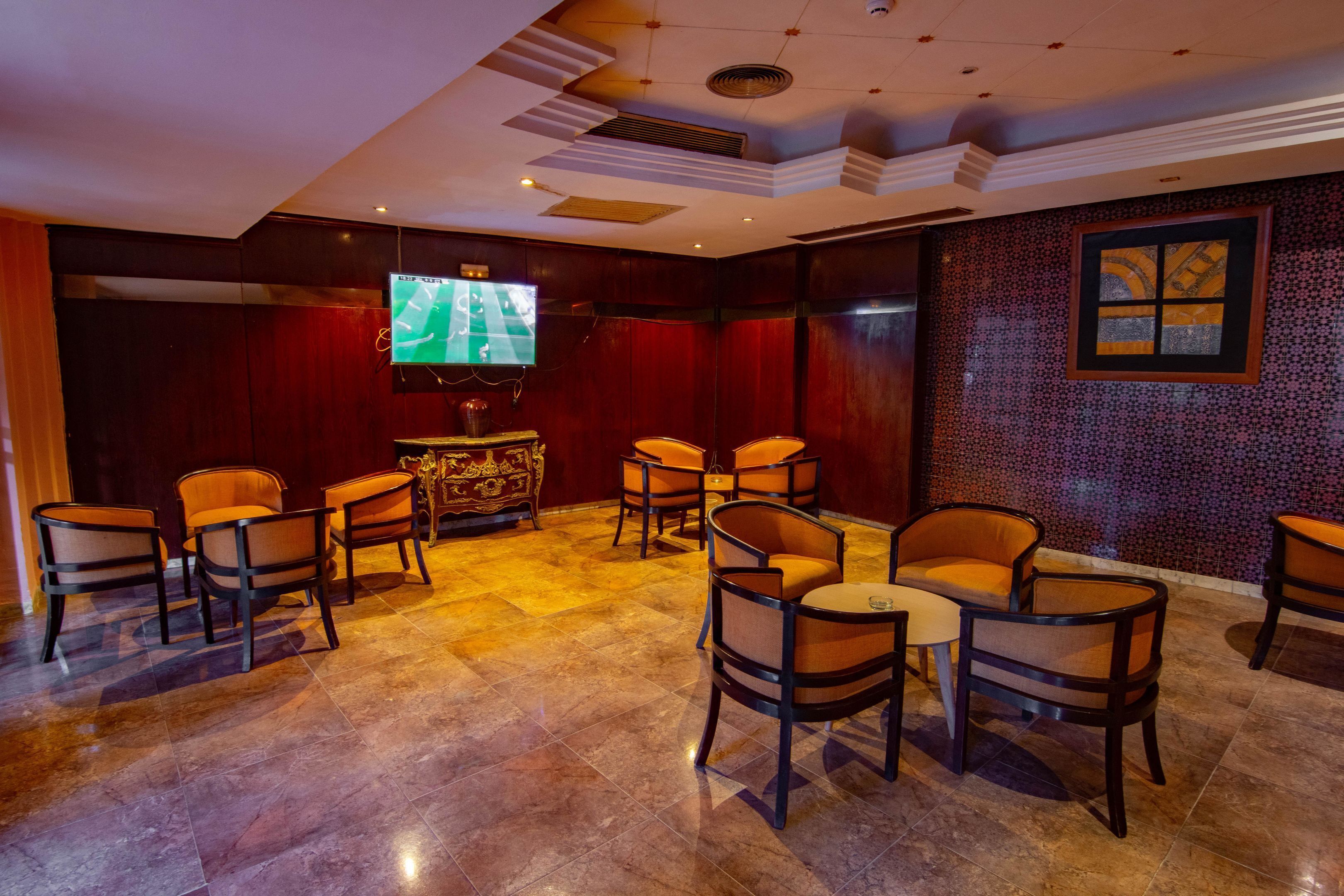 Een receptie met een groot scherm waarop een sportwedstrijd wordt getoond, verschillende zithoeken met stoelen en tafels, en een behang met patroon.