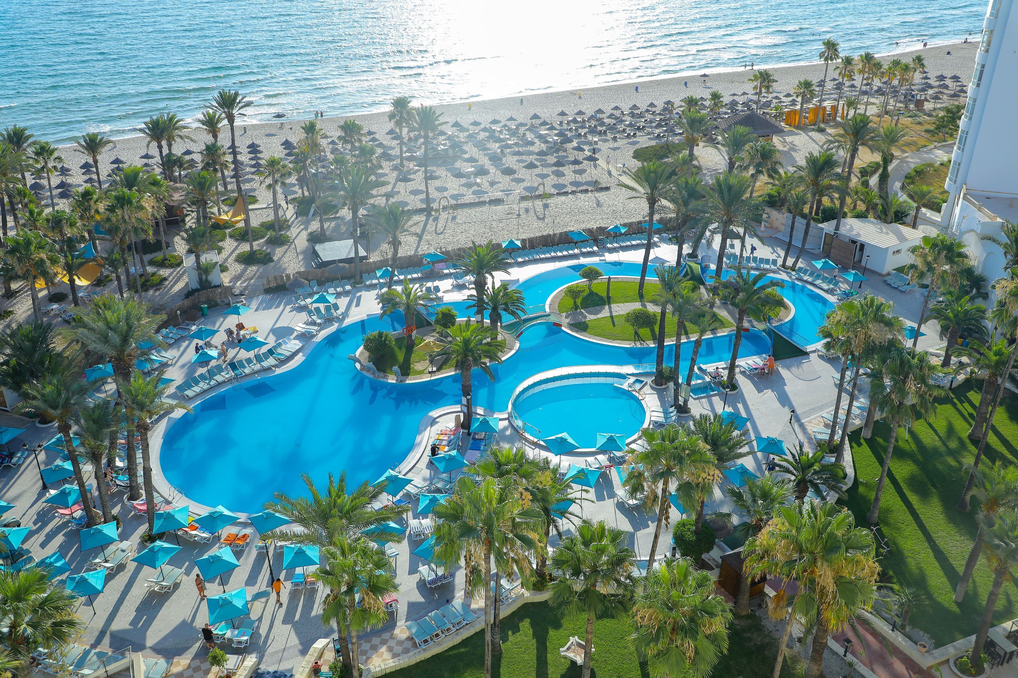 A resort with a large pool area, multiple sun loungers, and a beach with umbrellas.