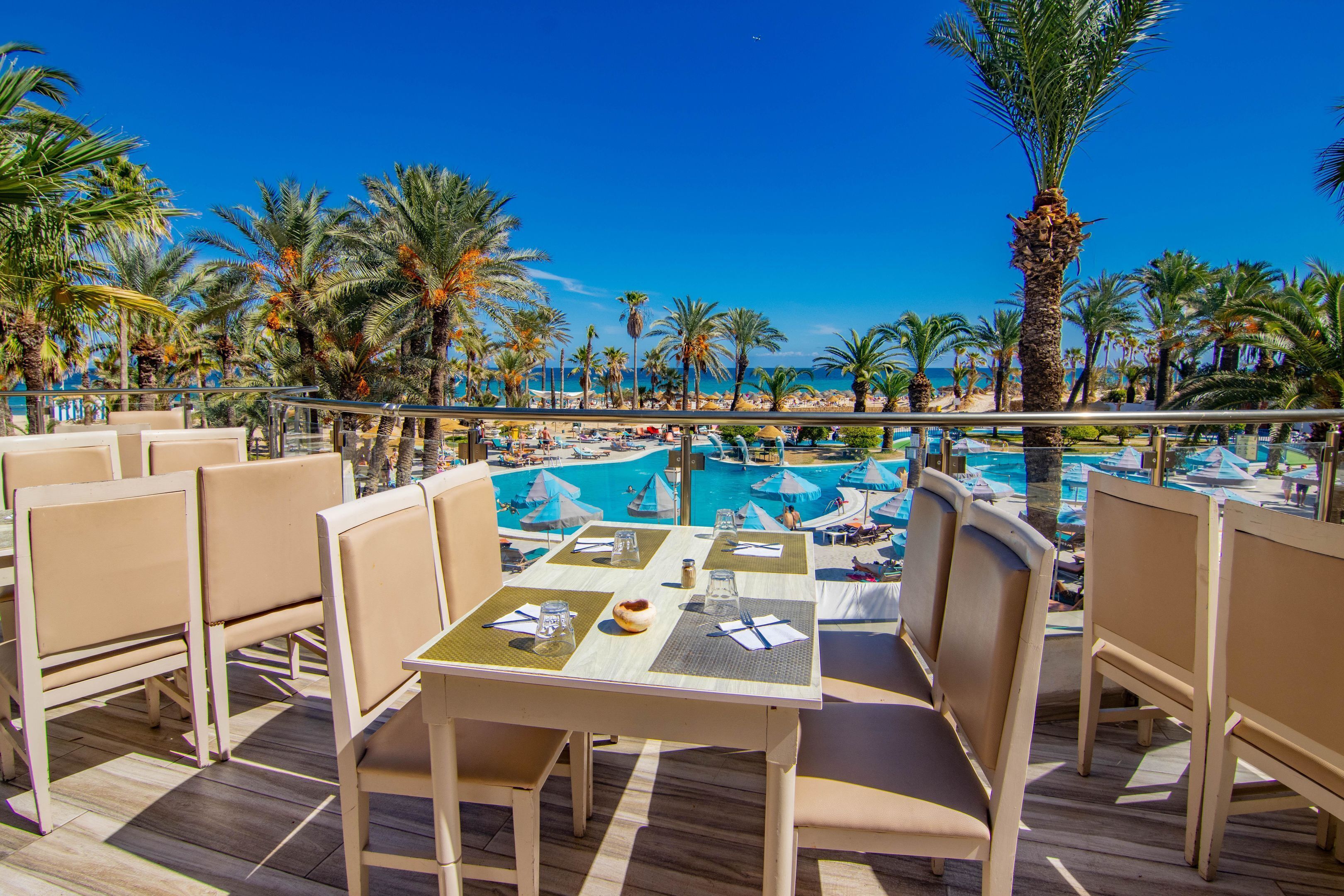 A poolside area with lounge chairs and tables, overlooking a pool and palm trees.