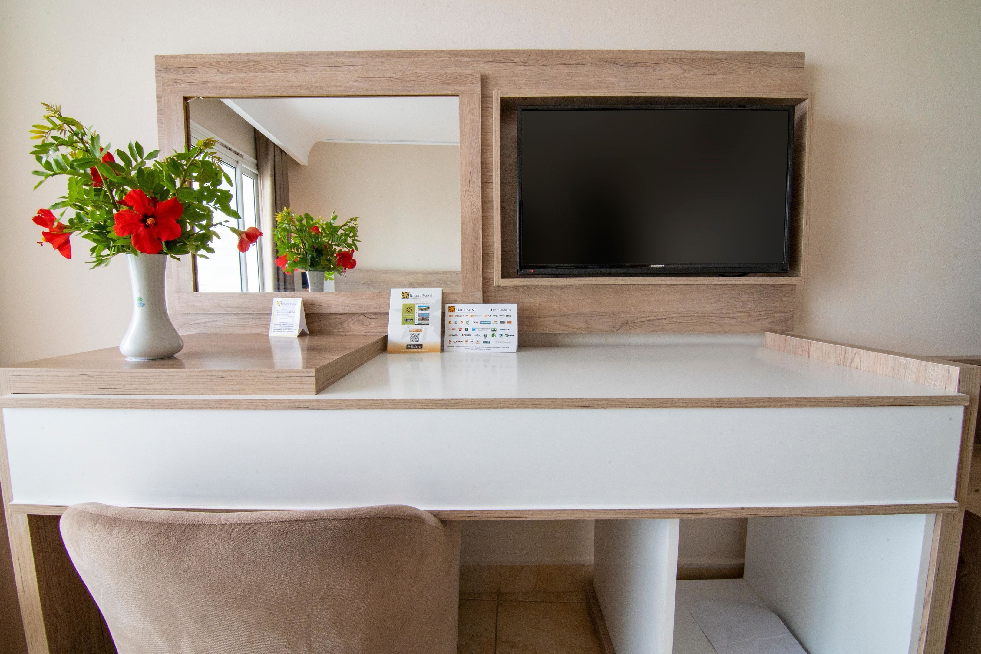 Habitació d'hotel moderna amb escriptori blanc, cadira beix, un mirall gran i televisió de pantalla plana penjada a la paret.