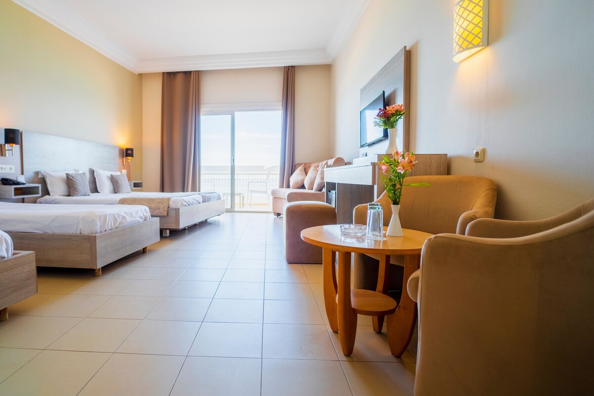 A hotel room with two beds, a sofa, a dining table, and a balcony with a view.
