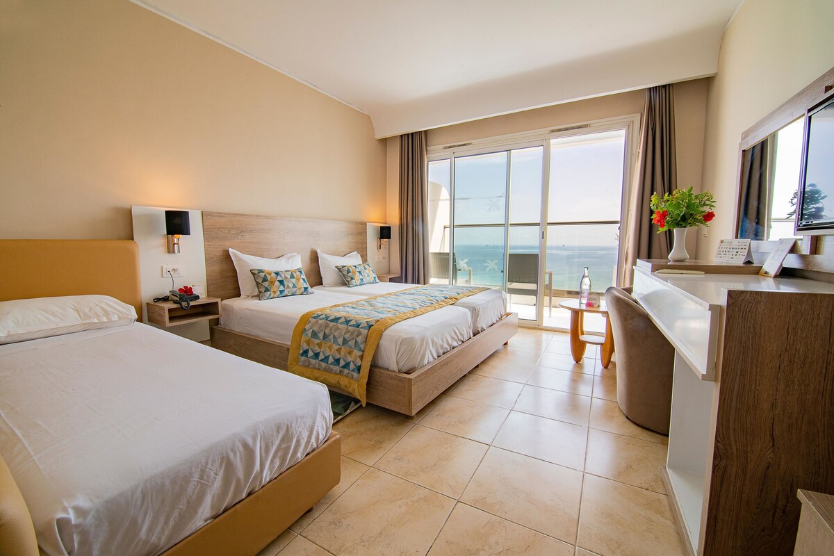 A hotel room with two beds, a desk, and a view of the sea.