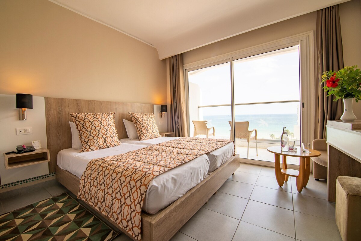 A hotel room with a large bed, a small table, a chair, a nightstand, and a view of the ocean.