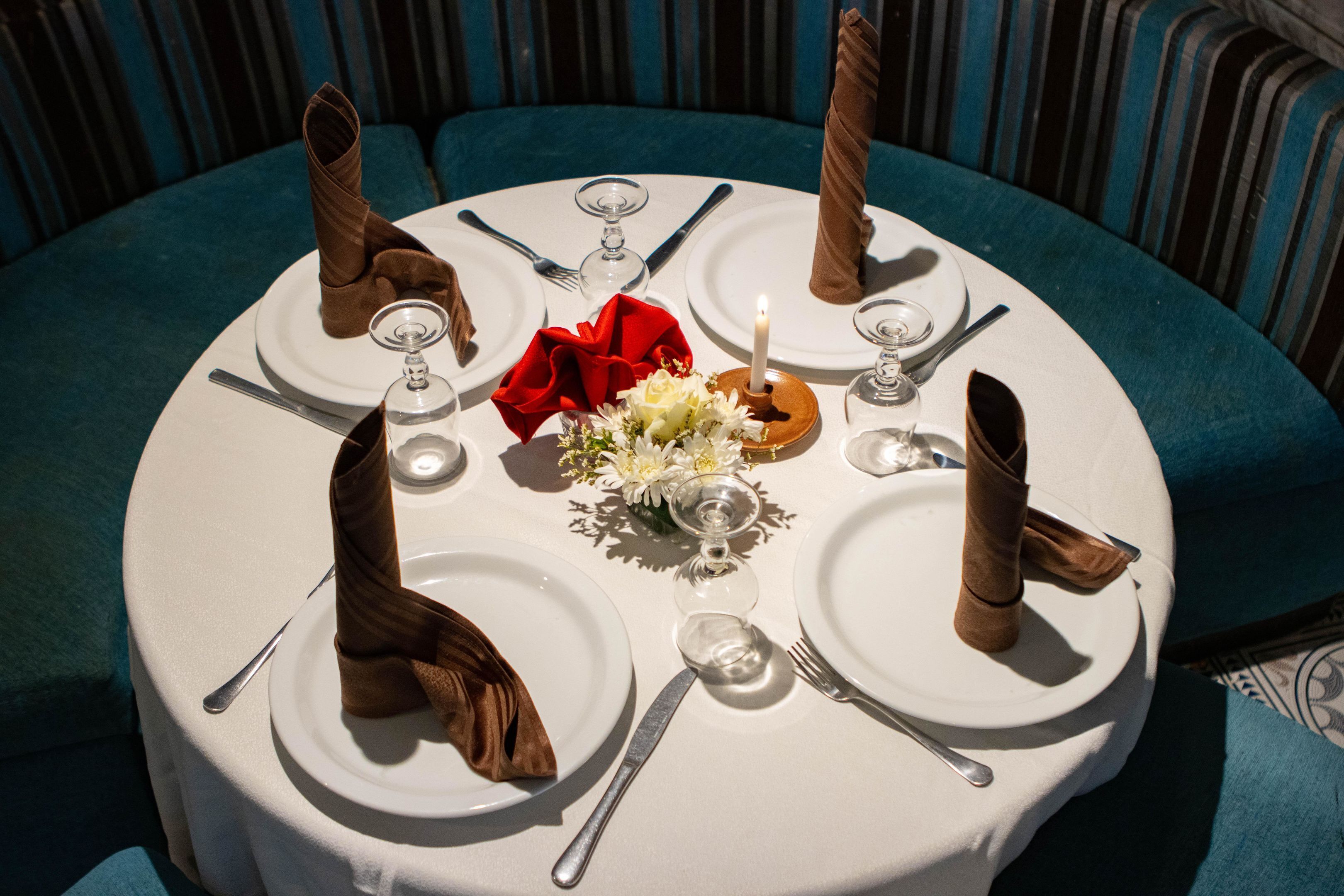 Une table ronde, dressée pour quatre personnes, avec de l’argenterie, des assiettes blanches et un centre de table composé de fleurs blanches et d’une serviette rouge.