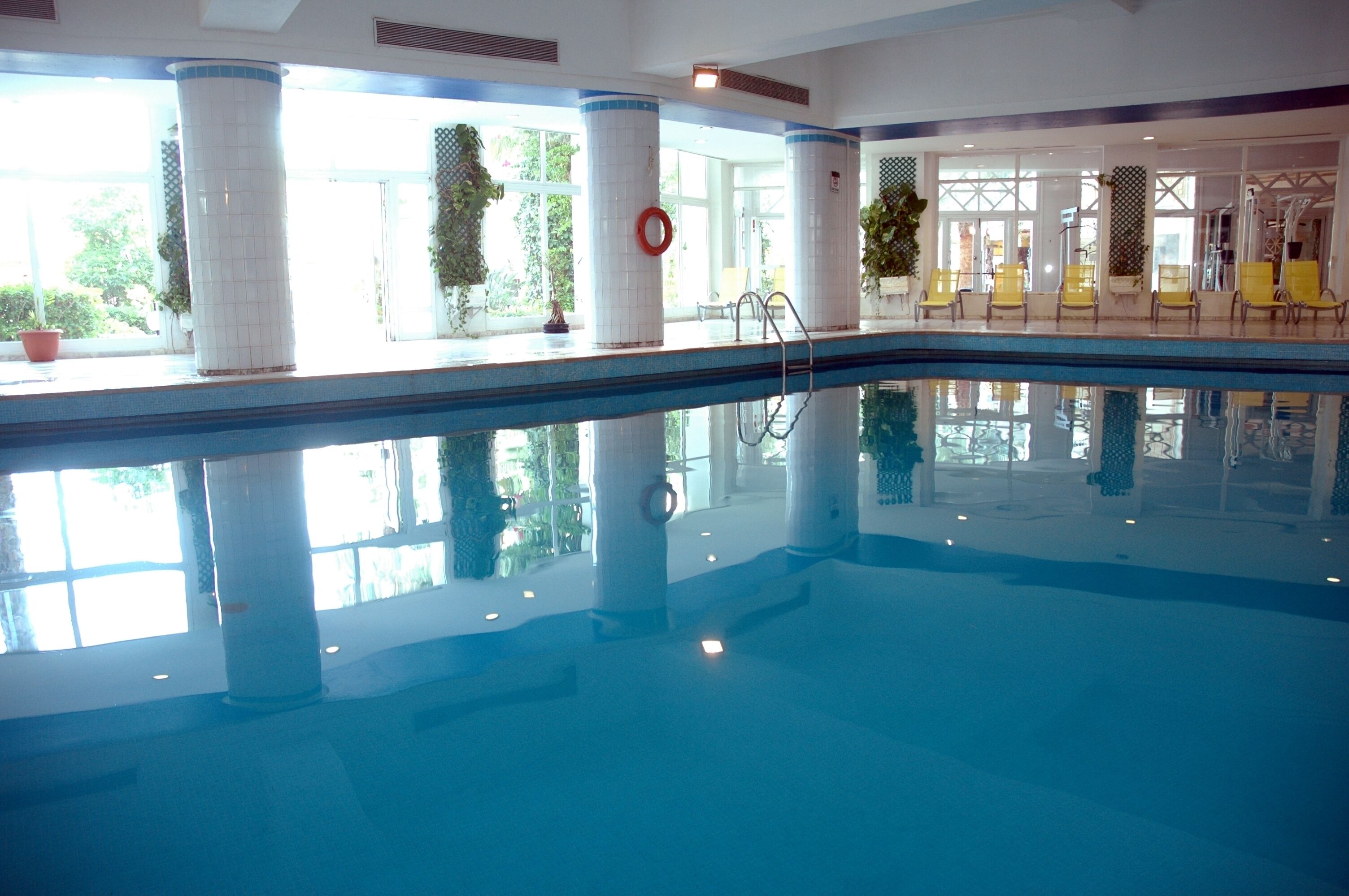 An indoor swimming pool with clear water, surrounded by white pillars, yellow chairs, and large windows allowing natural light.
