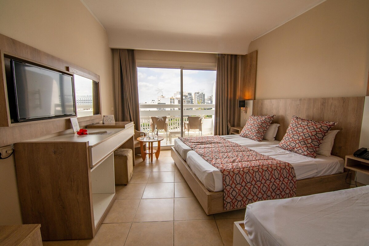 A hotel room with a balcony, a desk with a TV, two beds with patterned bedding, and a city view.