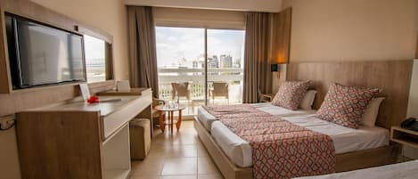 A hotel room with a balcony, a desk with a TV, two beds with patterned bedding, and a city view.