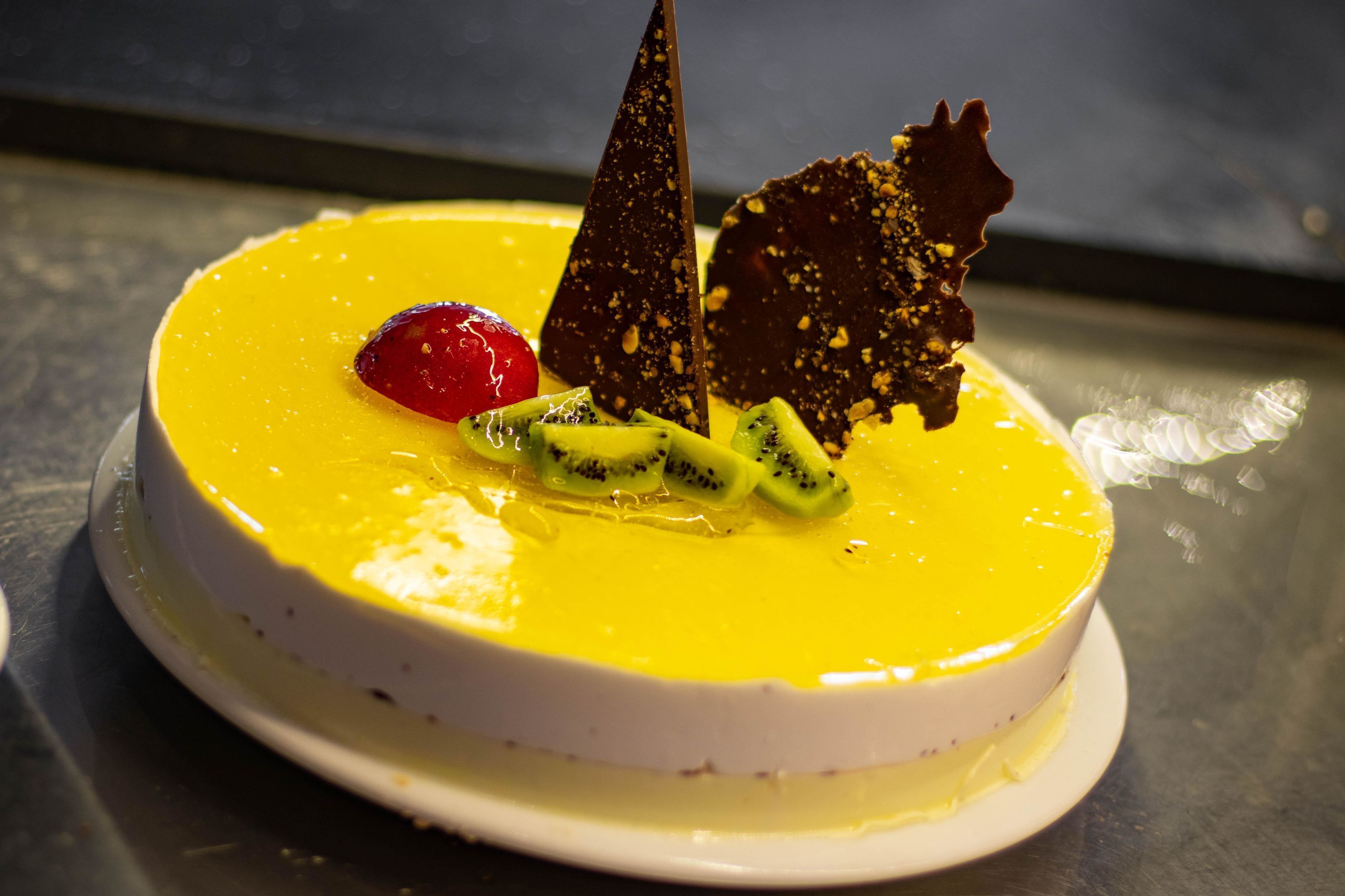 A round cake with yellow frosting, kiwi slices, a red cherry, and chocolate decorations on a white plate.