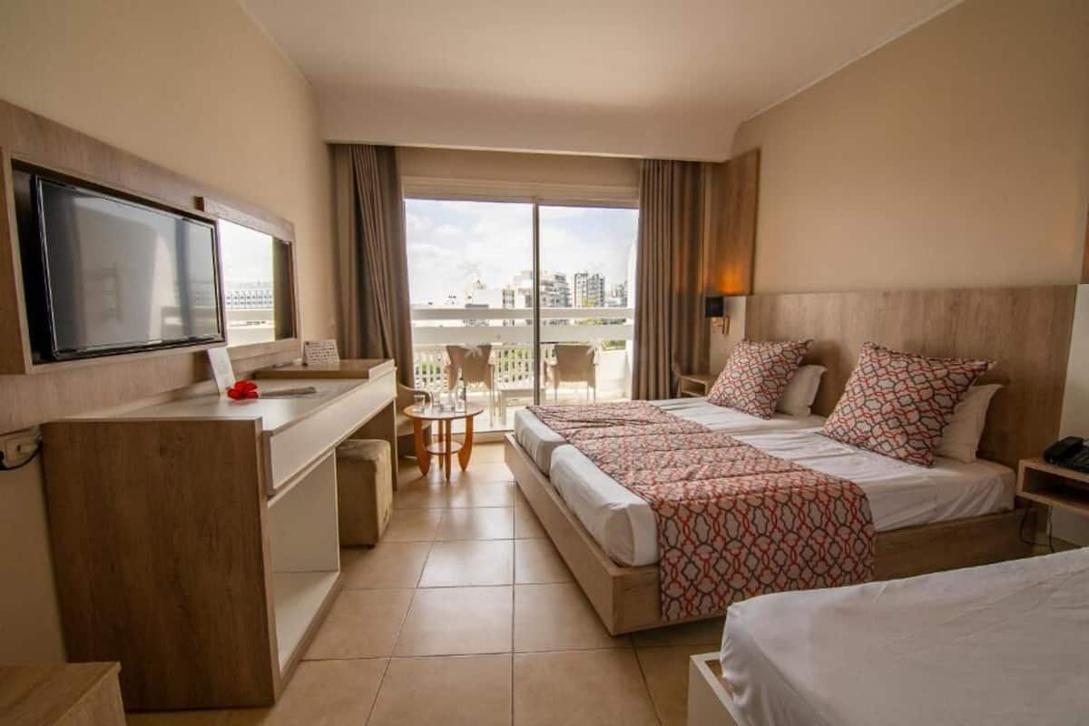A hotel room with a bed, a desk, a TV, and a balcony with a city view.