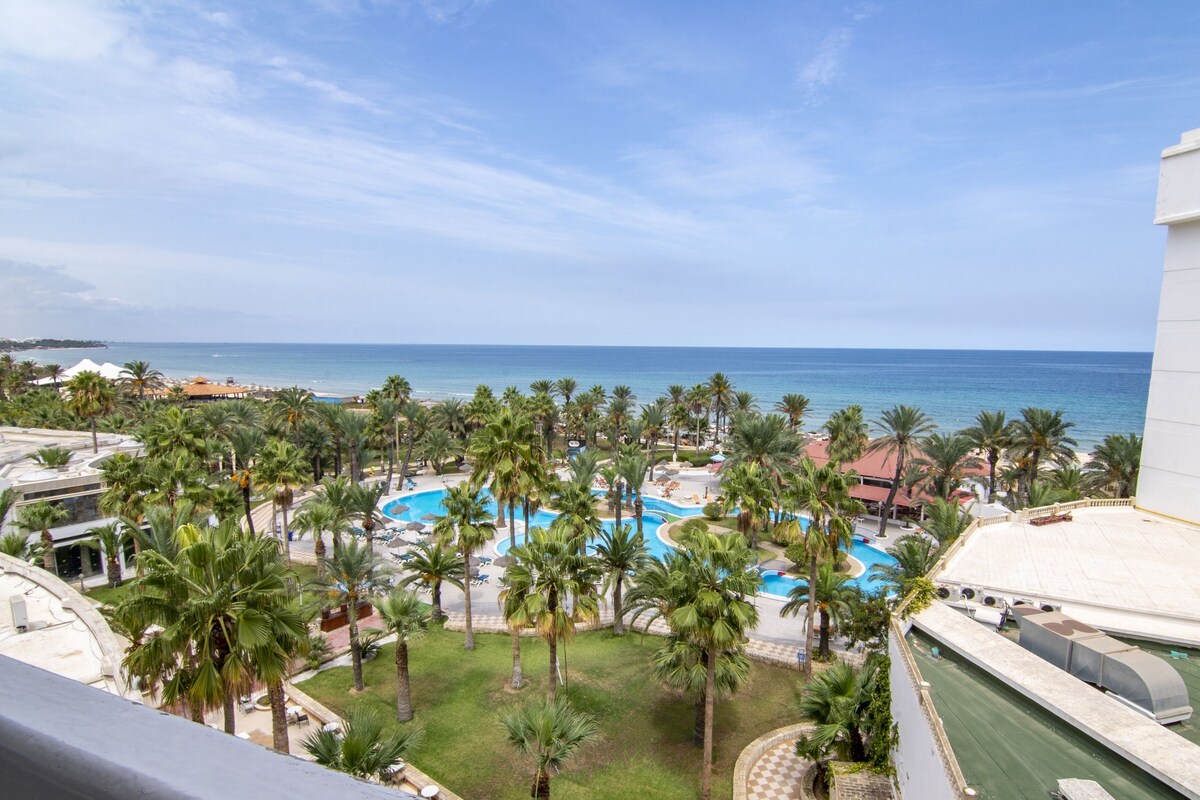 A resort with a pool surrounded by palm trees, a beach, and a view of the ocean.