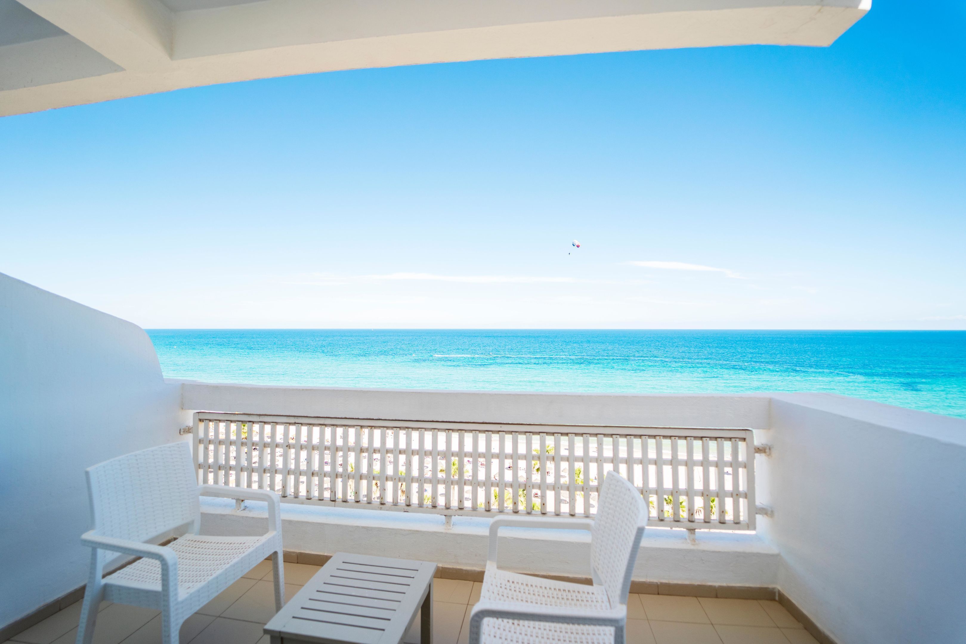 Ein Balkon mit zwei weißen Stühlen und einem Tisch, mit Blick auf einen Strand mit klarem, blauem Wasser und einem weißen Zaun.
