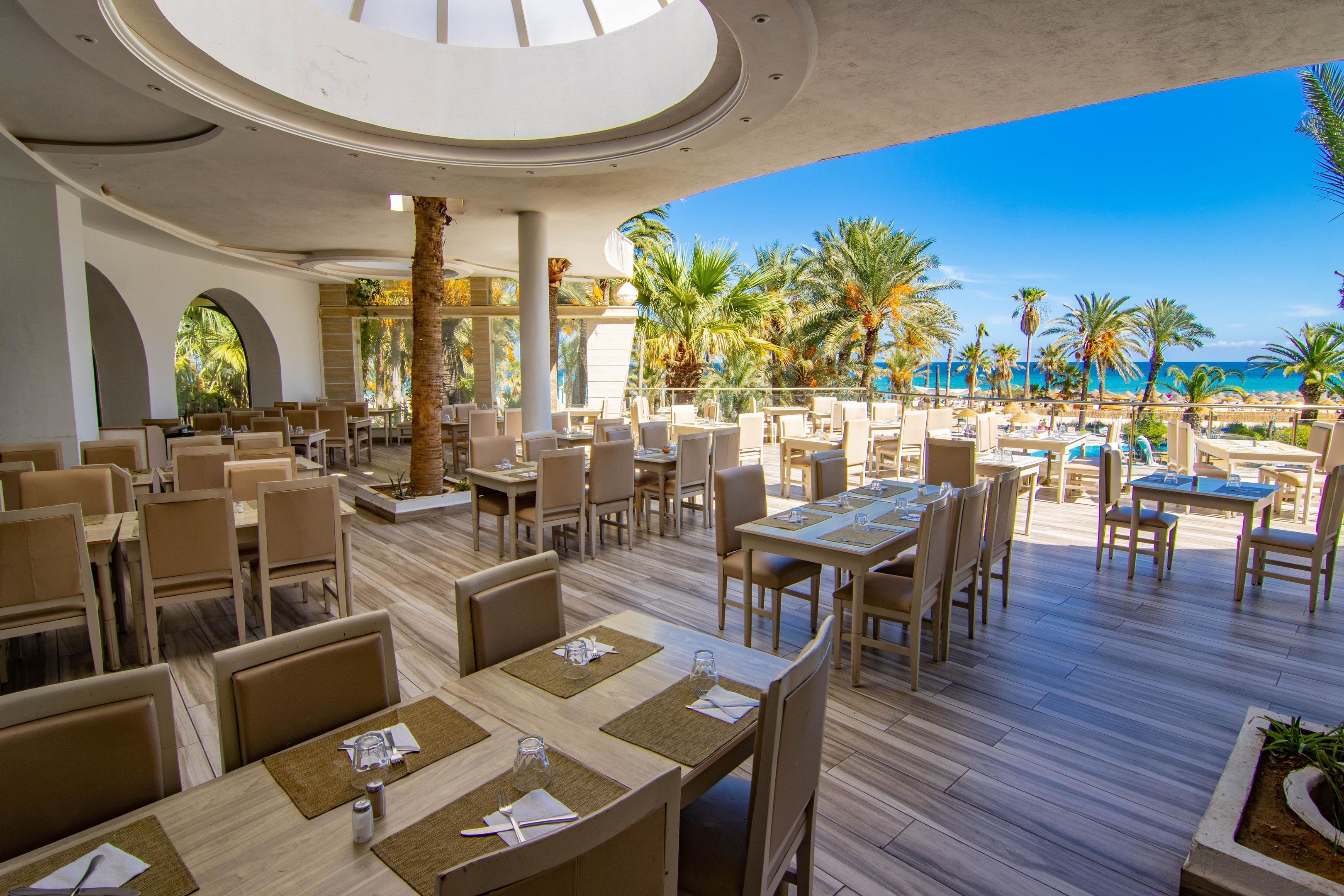 Un'ampia zona pranzo all'aperto con diversi tavoli e sedie, affacciata su una spiaggia con palme e l'oceano.