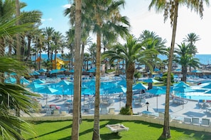 A pool area with multiple blue umbrellas and lounge chairs, surrounded by palm trees. - Riadh Palms Resort & Spa (Sousse)