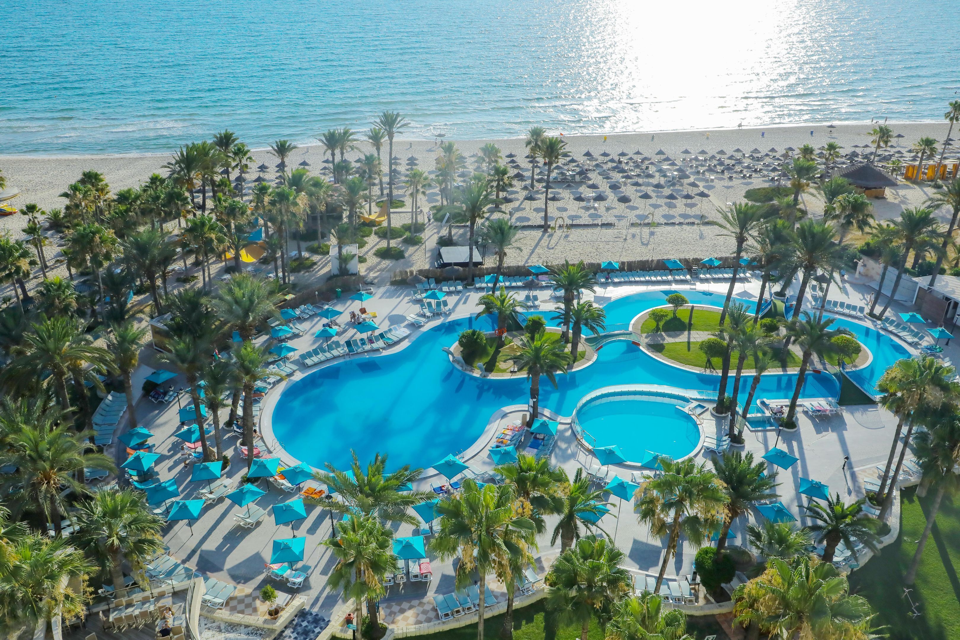 A resort with a central swimming pool, multiple lounge areas, and a beach with umbrellas.