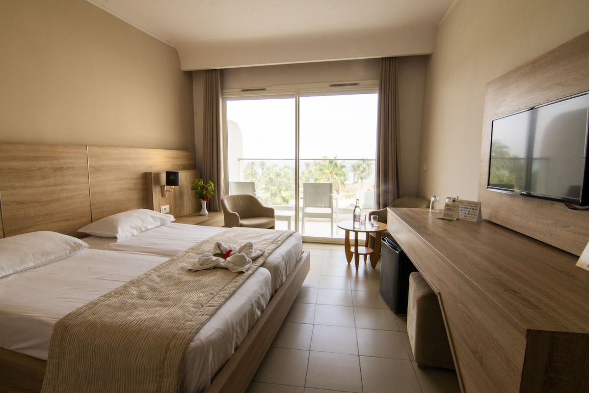 A hotel room with two beds, a desk, a chair, a TV, and a balcony with a view.