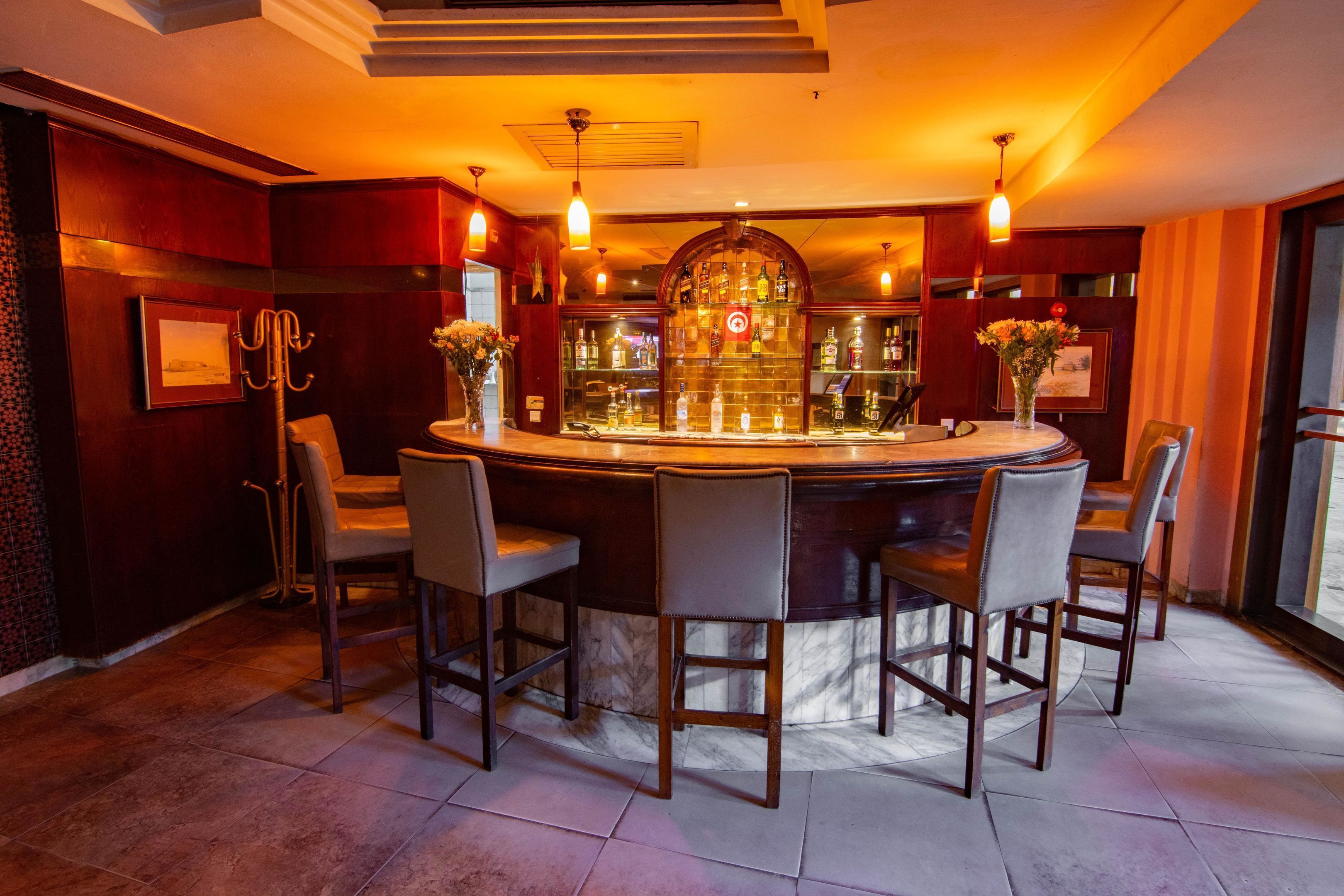 Eine Bar mit einem runden Tisch und vier Stühlen, einer Theke voller Flaschen und einer Wand mit gerahmten Bildern sowie einem Blumenarrangement.