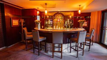 Eine Bar mit einem runden Tisch und vier Stühlen, einer Theke voller Flaschen und einer Wand mit gerahmten Bildern sowie einem Blumenarrangement.