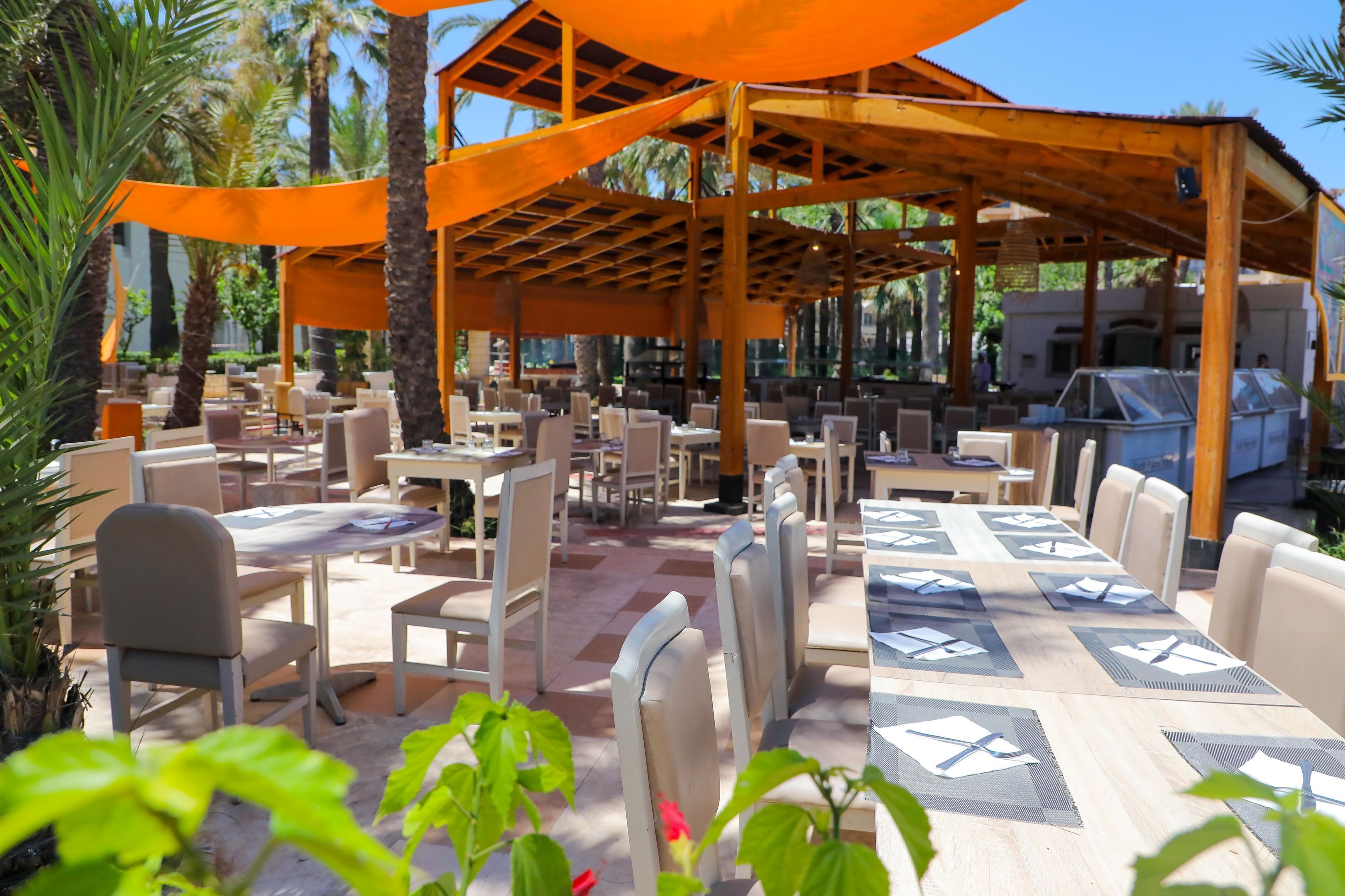 An outdoor restaurant with tables and chairs, covered by a wooden pergola, surrounded by palm trees.