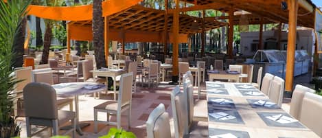 An outdoor restaurant with tables and chairs, covered by a wooden pergola, surrounded by palm trees.