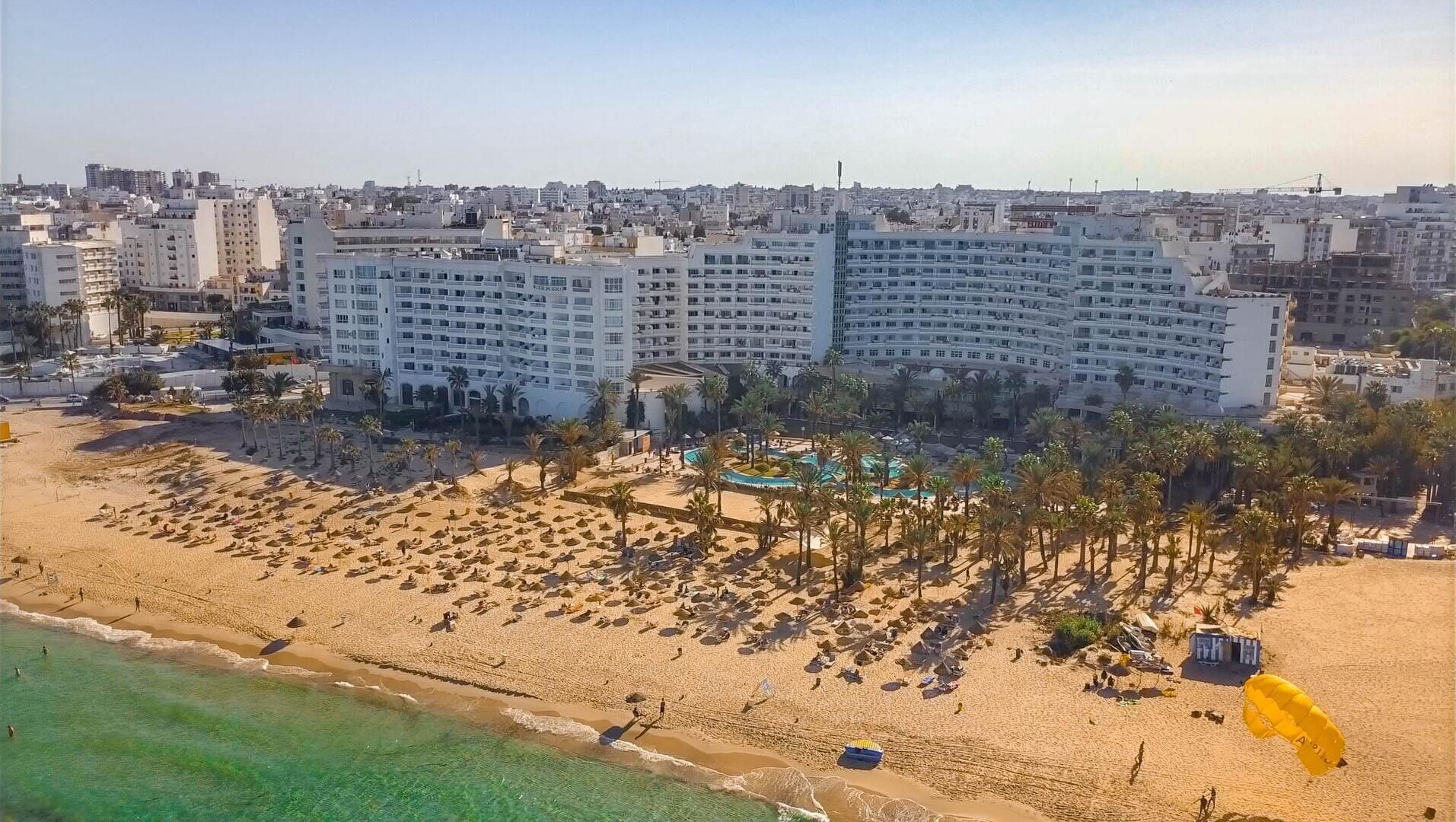 A large hotel with multiple floors, a beach area with sun loungers and umbrellas, and a pool.