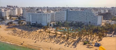A large hotel with multiple floors, a beach area with sun loungers and umbrellas, and a pool.