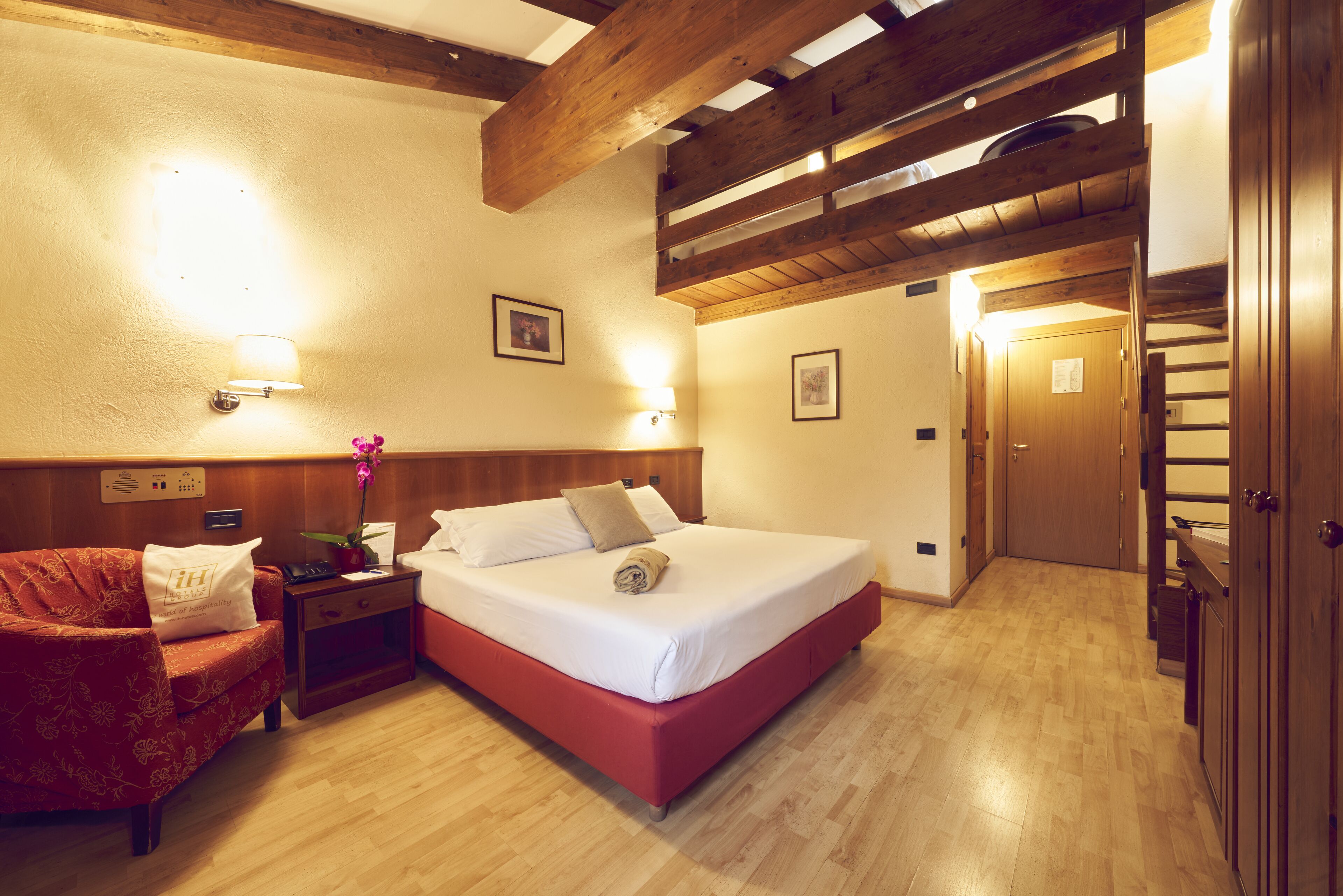 A hotel room with a wooden ceiling, a bed with white bedding, a red sofa, a nightstand with a lamp, and a wooden wardrobe.