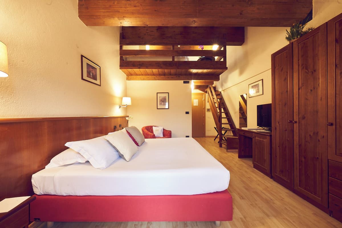 A hotel room with a wooden headboard, a bed with white bedding, a red accent on the bed, a wooden wardrobe, a desk with a computer, and a staircase.