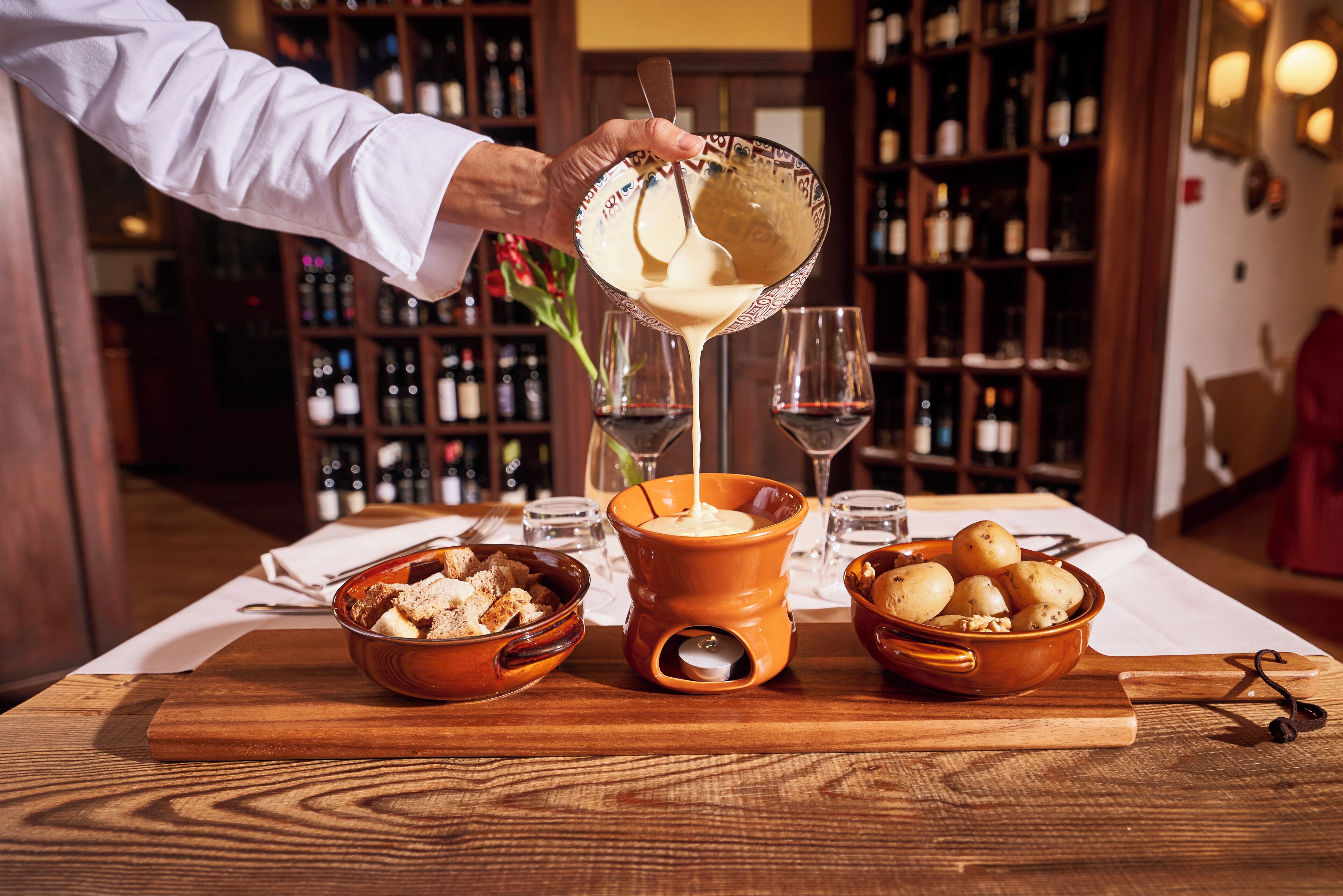 A person pouring a creamy substance into a bowl, with wine glasses and a wooden table in the background.