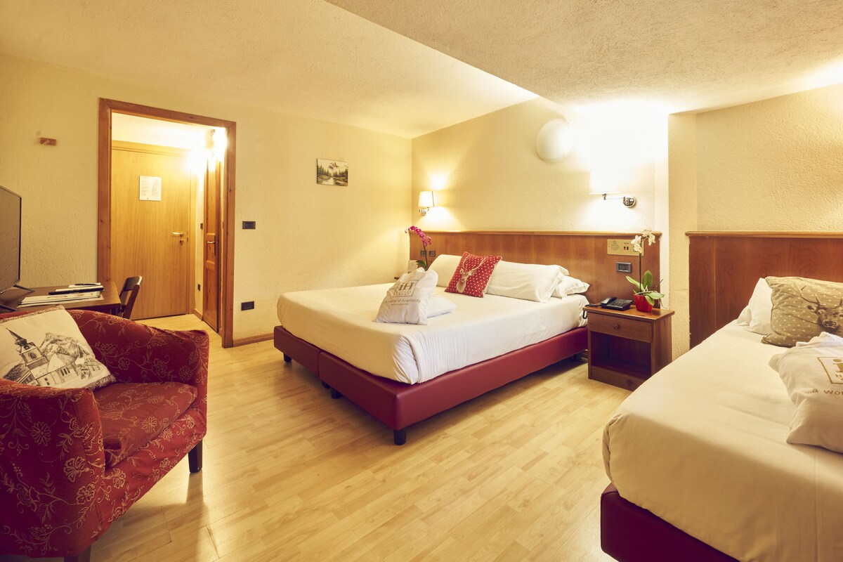 A hotel room with a large bed, a smaller bed, a red armchair, a wooden nightstand, and a desk with a lamp.