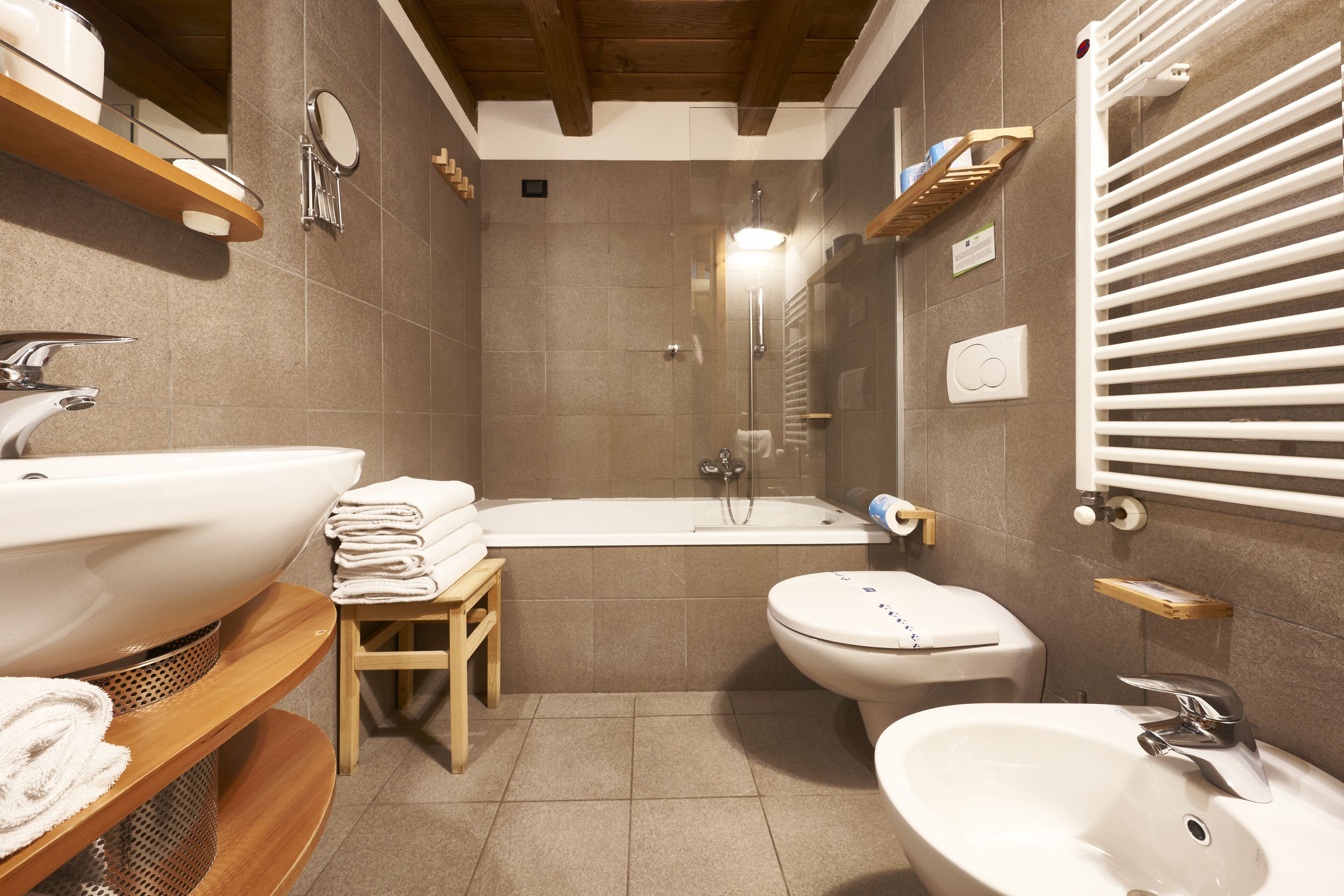 A modern bathroom with a large sink, toilet, and bathtub. There are wooden shelves with towels and toiletries.