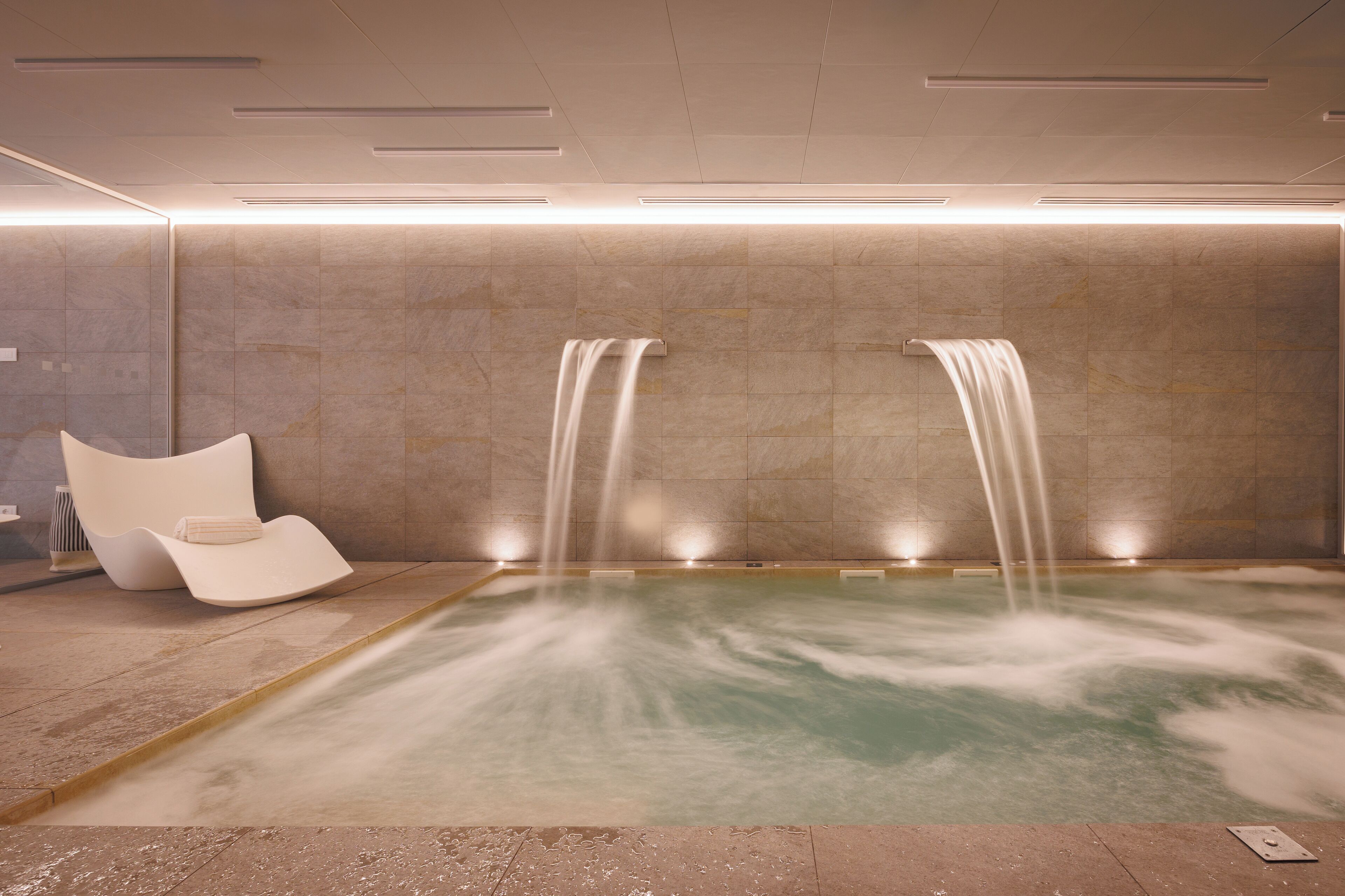 A spa room with a hot tub, two waterfalls, and a modern chair.