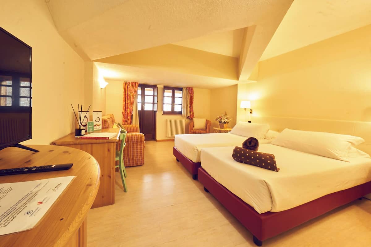 A hotel room with a desk, two beds, a chair, a window with curtains, and a radiator.