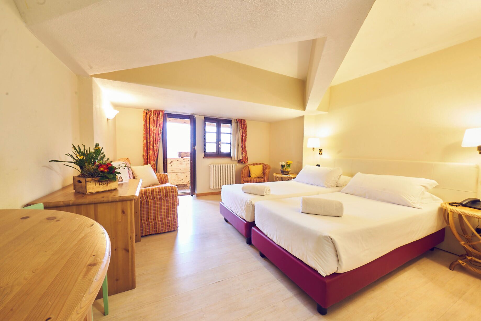 Camera d'albergo con due letti, una scrivania in legno, una sedia, un tavolino con una pianta e vista all'esterno tramite una porta.